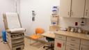 A clinical examination room with a brown examination table, orange chair, medical equipment on the wall, and a cabinet holding medical supplies. A clinical examination room with a brown examination table, orange chair, medical equipment on the wall, and a cabinet holding medical supplies.