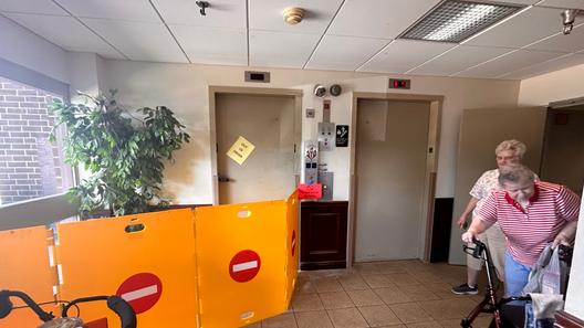 Two women, one leaning on a walker, stand in a hall near two elevators. One elevator with an out of order sign on the door has four barriers in front of it. Two women, one leaning on a walker, stand in a hall near two elevators. One elevator with an out of order sign on the door has four barriers in front of it.