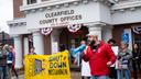 A man in a red jacket speaks into a megaphone in front of the Clearfield County Offices building. Next to him are protesters holding a yellow sign that says “Shut Down Moshannon.” A man in a red jacket speaks into a megaphone in front of the Clearfield County Offices building. Next to him are protesters holding a yellow sign that says “Shut Down Moshannon.”