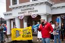 A man in a red jacket speaks into a megaphone in front of the Clearfield County Offices building. Next to him are protesters holding a yellow sign that says “Shut Down Moshannon.” A man in a red jacket speaks into a megaphone in front of the Clearfield County Offices building. Next to him are protesters holding a yellow sign that says “Shut Down Moshannon.”