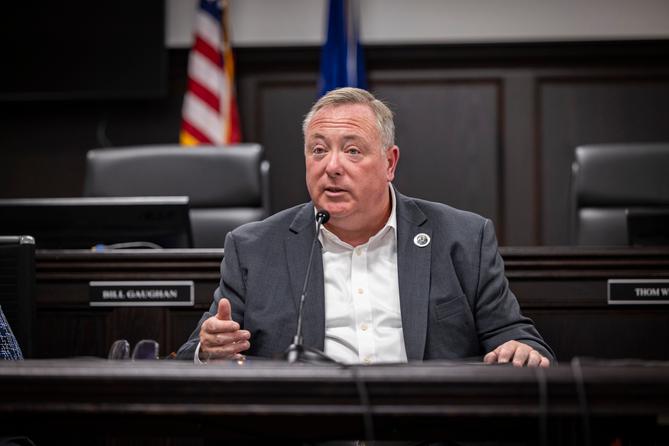 A man identified as Aging Secretary Jason Kavulich sits at a desk in a dark wood-paneled government hearing room. A man identified as Aging Secretary Jason Kavulich sits at a desk in a dark wood-paneled government hearing room.